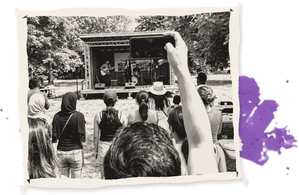 A crowd in a park watching a small open-air stage, one phone held up